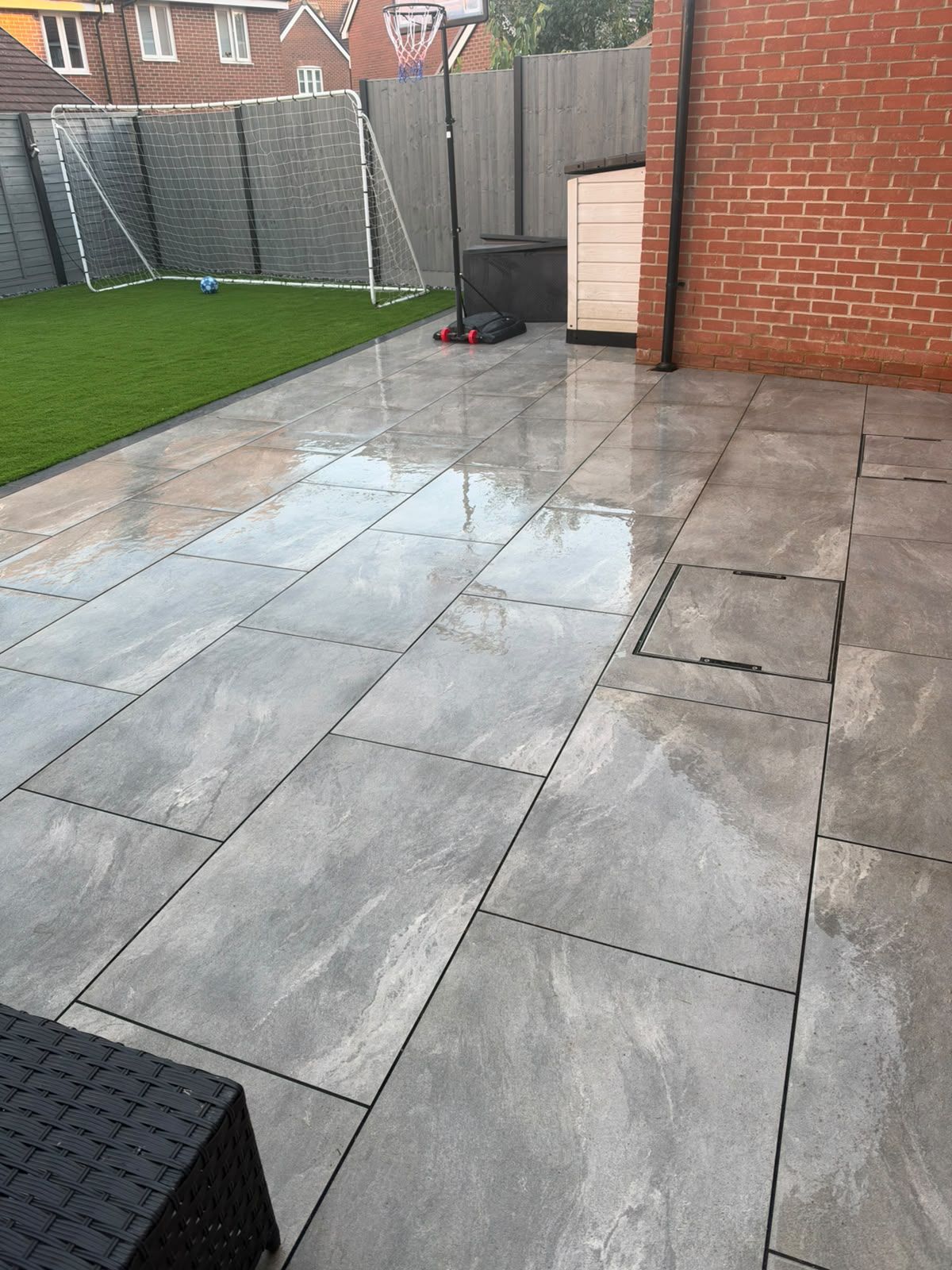Progreen-installed grey patio paving meeting artificial grass beside a brick wall and access cover