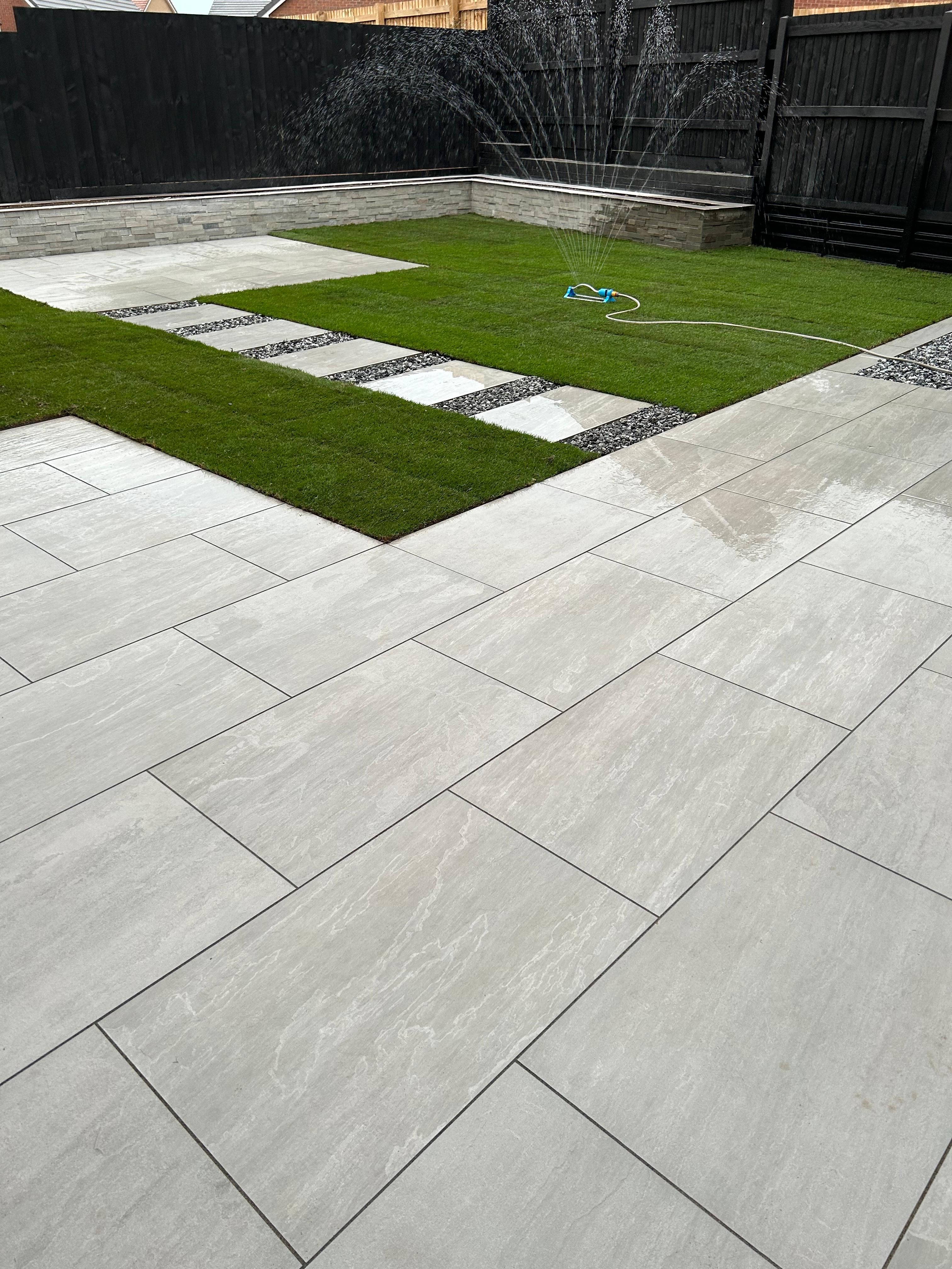A garden with pale paving, stepping-stone path, green turf and dark timber boundary photographed during watering